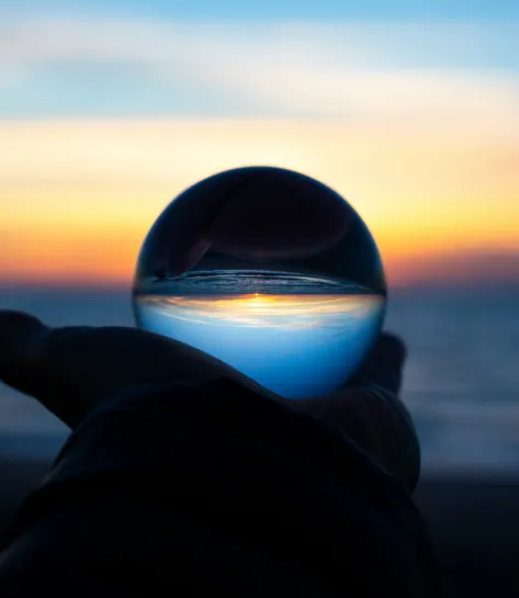A hand holding a glass sphere that reflects an inverted seaside sunset.