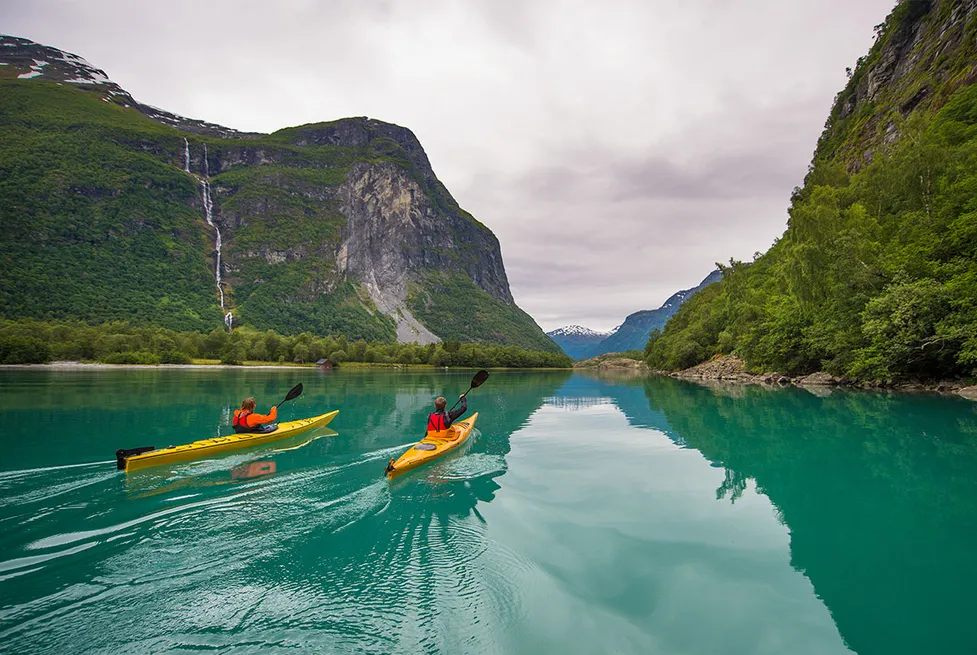 Two people kayaking on a turquoise fjord lake surrounded by steep mountains and a waterfall in Stryn, Norway.
