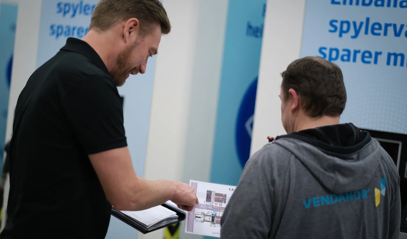 Two Vendanor employees review a printed product drawing together in front of branded exhibition banners.