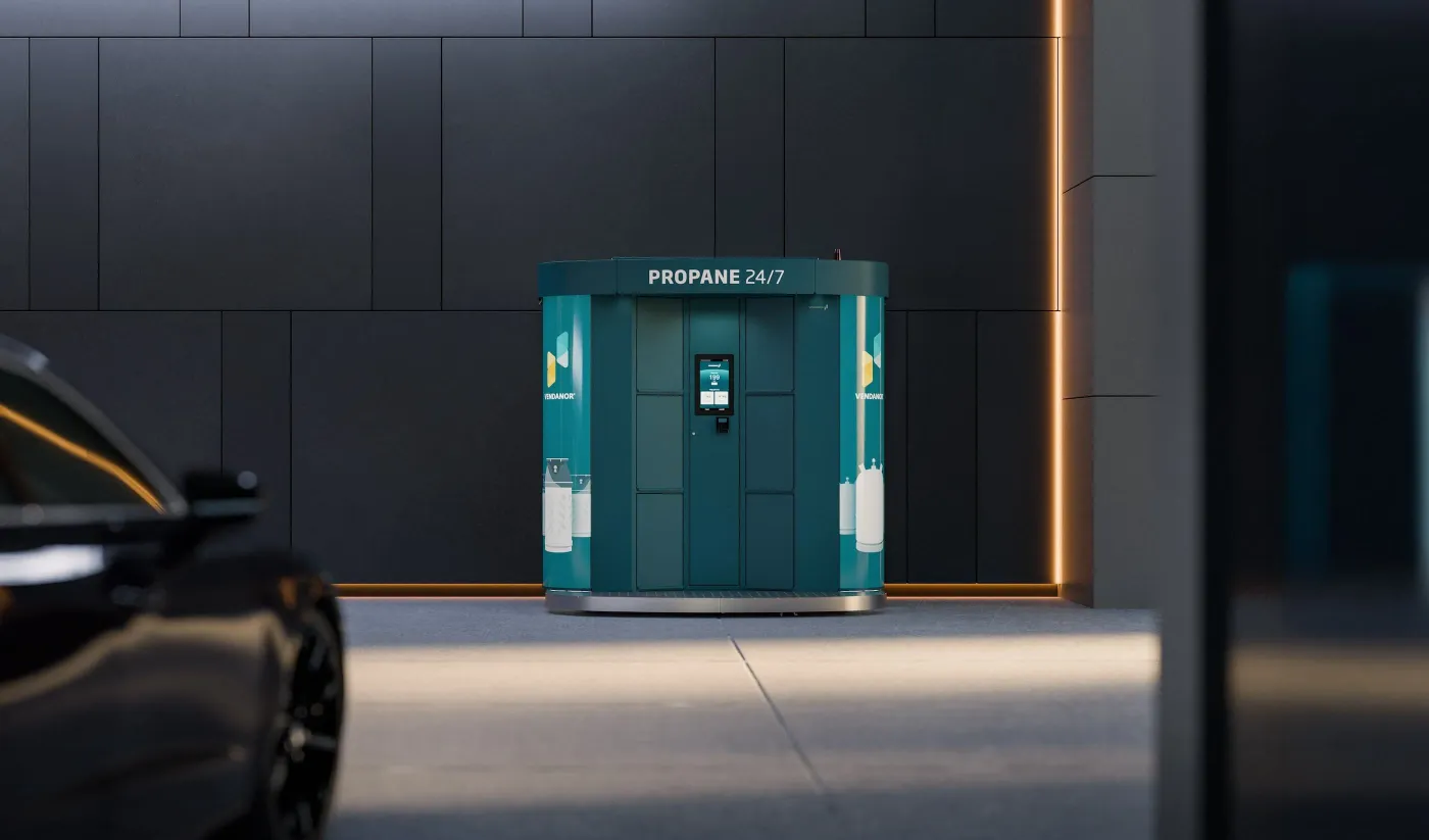 A teal Vendanor Propane Vending Machine labelled Propane 24/7 stands against a dark panelled wall with a car in the foreground.