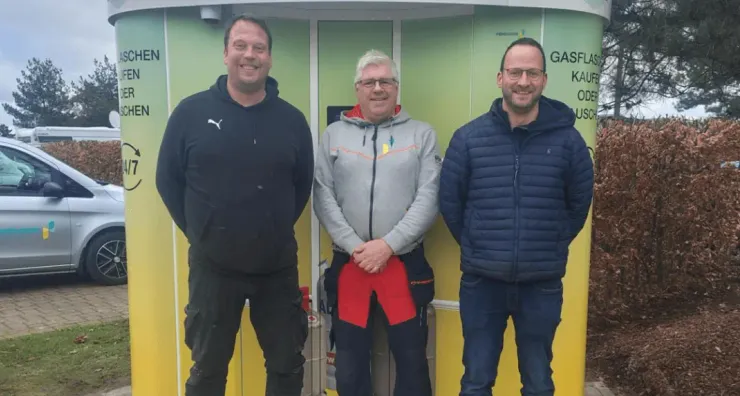 Three men standing in front of a green and yellow Vendanor Propane Vending Machine at Emsland camp.