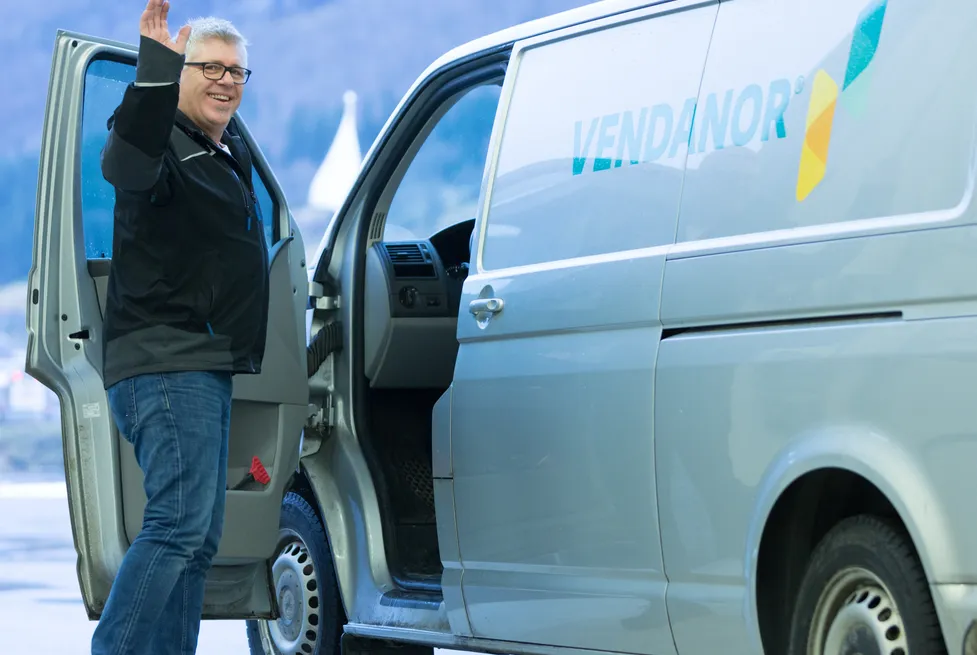 A smiling Vendanor service technician waving beside a branded Vendanor service van in a Norwegian mountain setting.