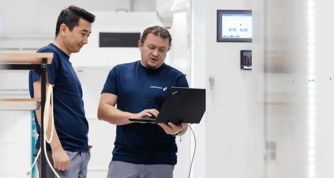 Two Vendanor technicians in branded T-shirts testing a machine with a laptop in the production facility.